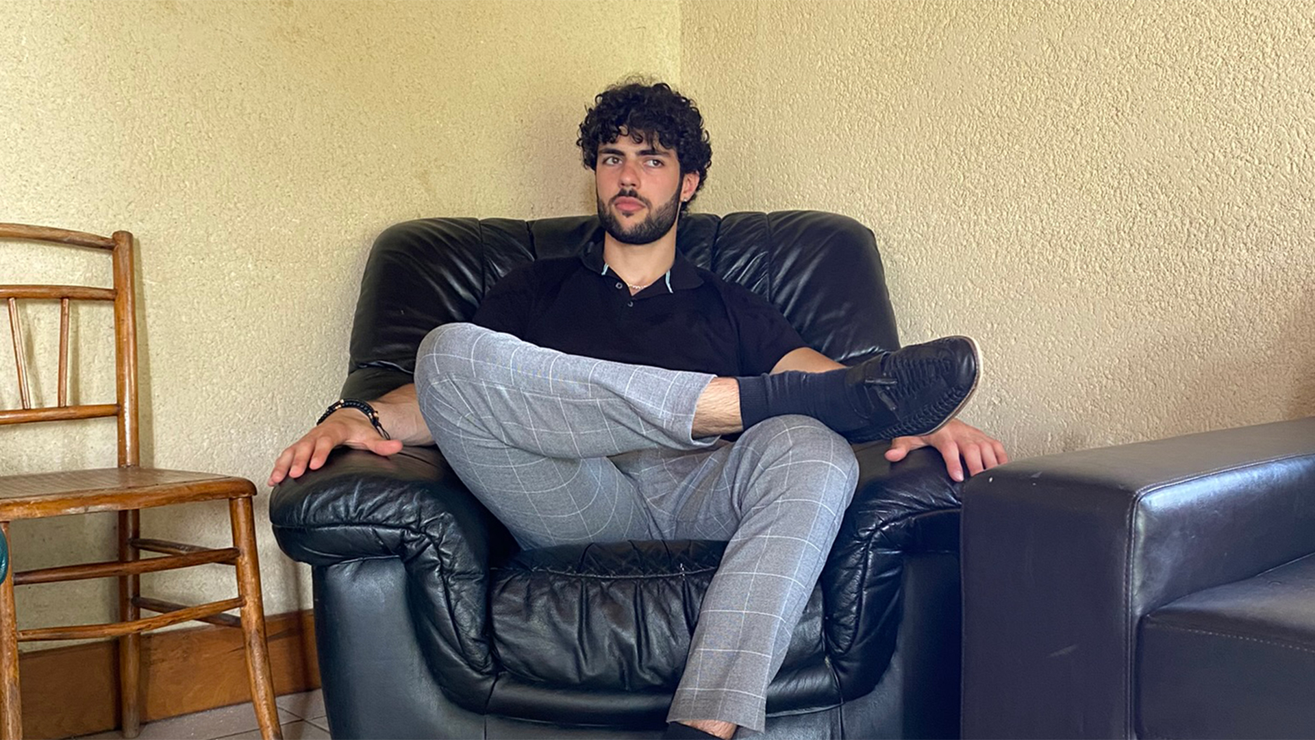 Matteo sitting on a leather coach with a black top and grey trousers with his legs crossed.